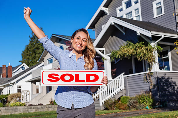 Photo of a cheerful young Hispanic businesswoman/realtor with a red SOLD sign, standing in front of a row of suburban houses.
